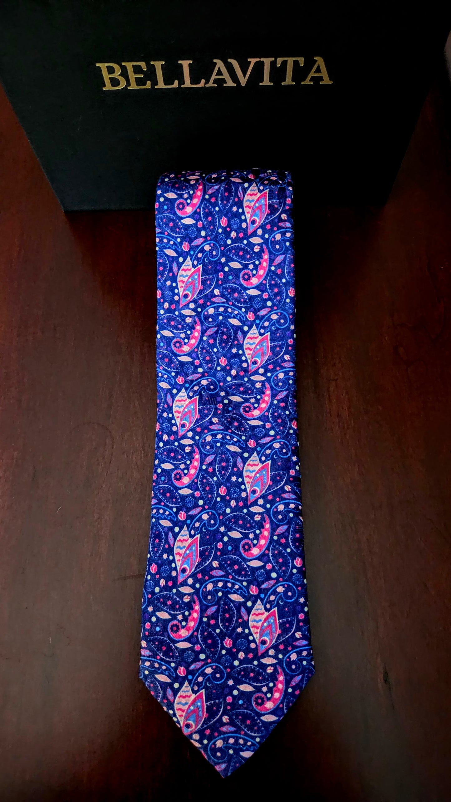 A blue paisley necktie with pink and white patterns, displayed vertically in front of a black Bellavita box on a dark wooden surface.