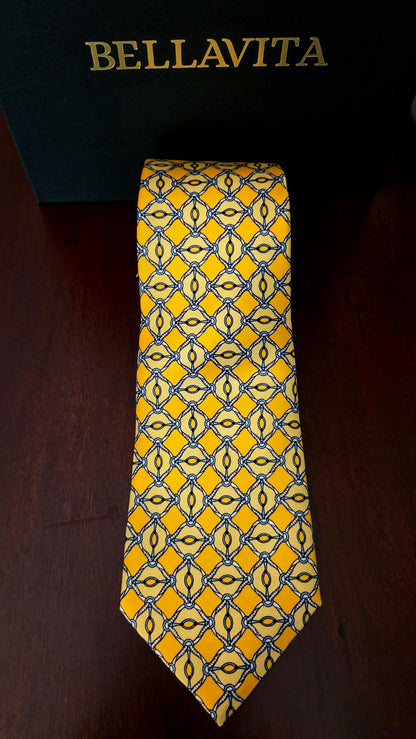 A yellow Bellavita necktie with a blue and black geometric pattern, displayed in front of its black branded box on a dark wooden surface.
