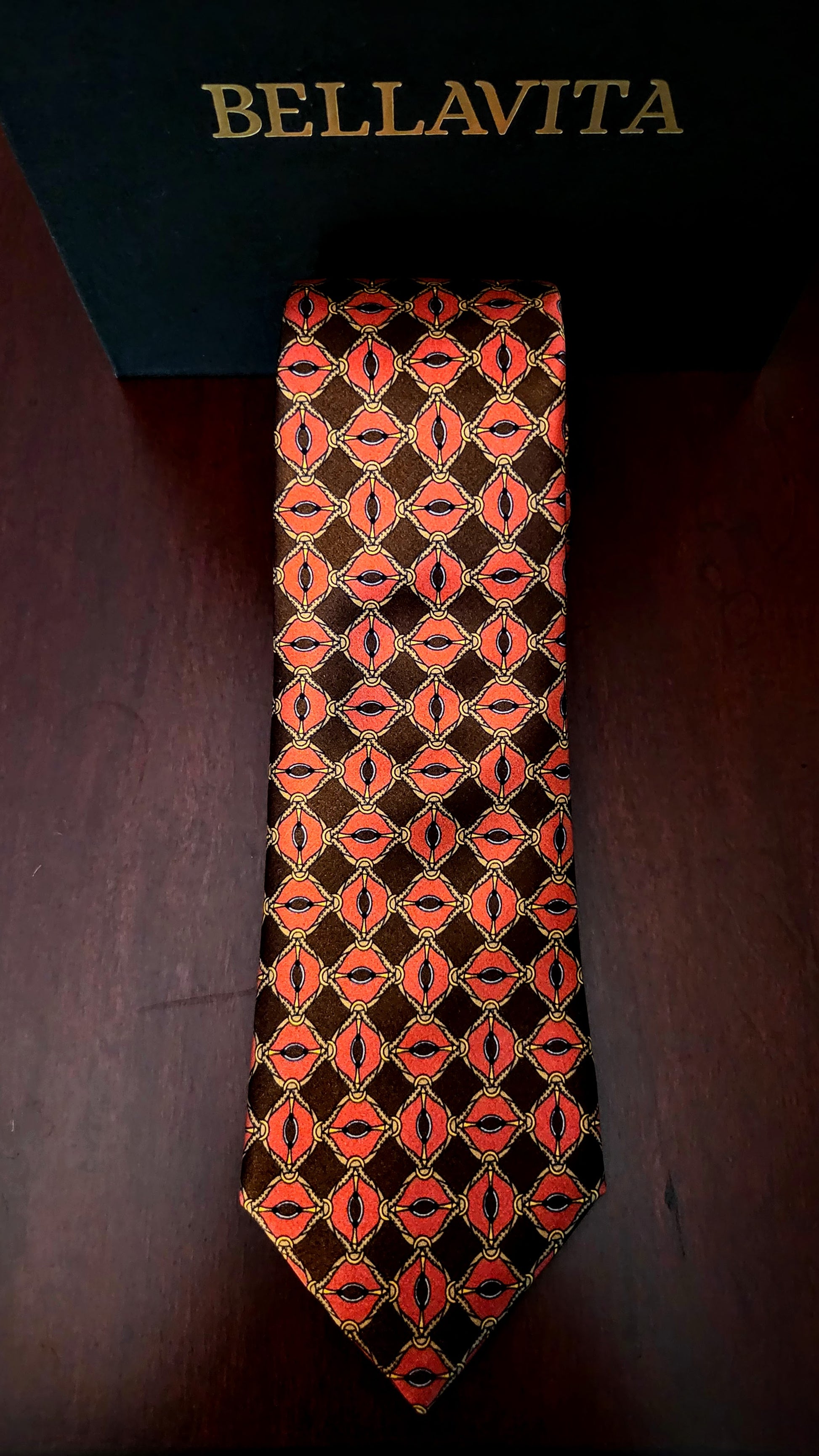A Bellavita necktie with a repeating geometric pattern of red and orange oval motifs on a dark brown background, displayed vertically on a dark wooden surface with its black branded box visible at the top.