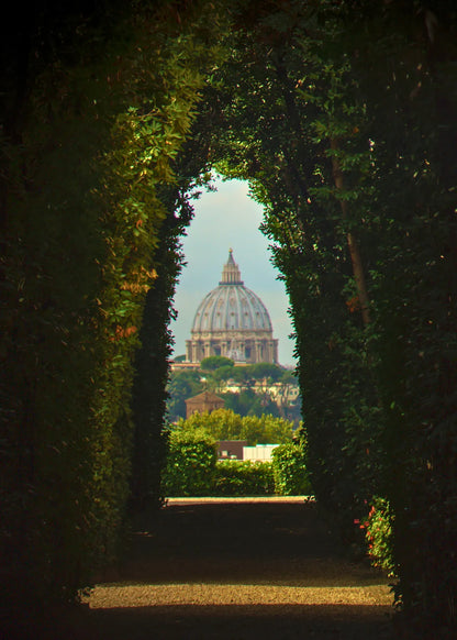 A scenic view of St. Peter's dome in Vatican City framed by a dark, leafy hedge archway in the foreground.