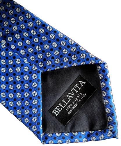 A close-up of a blue necktie with a repeating geometric pattern of white and black shapes, featuring a black fabric brand label sewn onto the inside that reads 'BELLA VITA'.