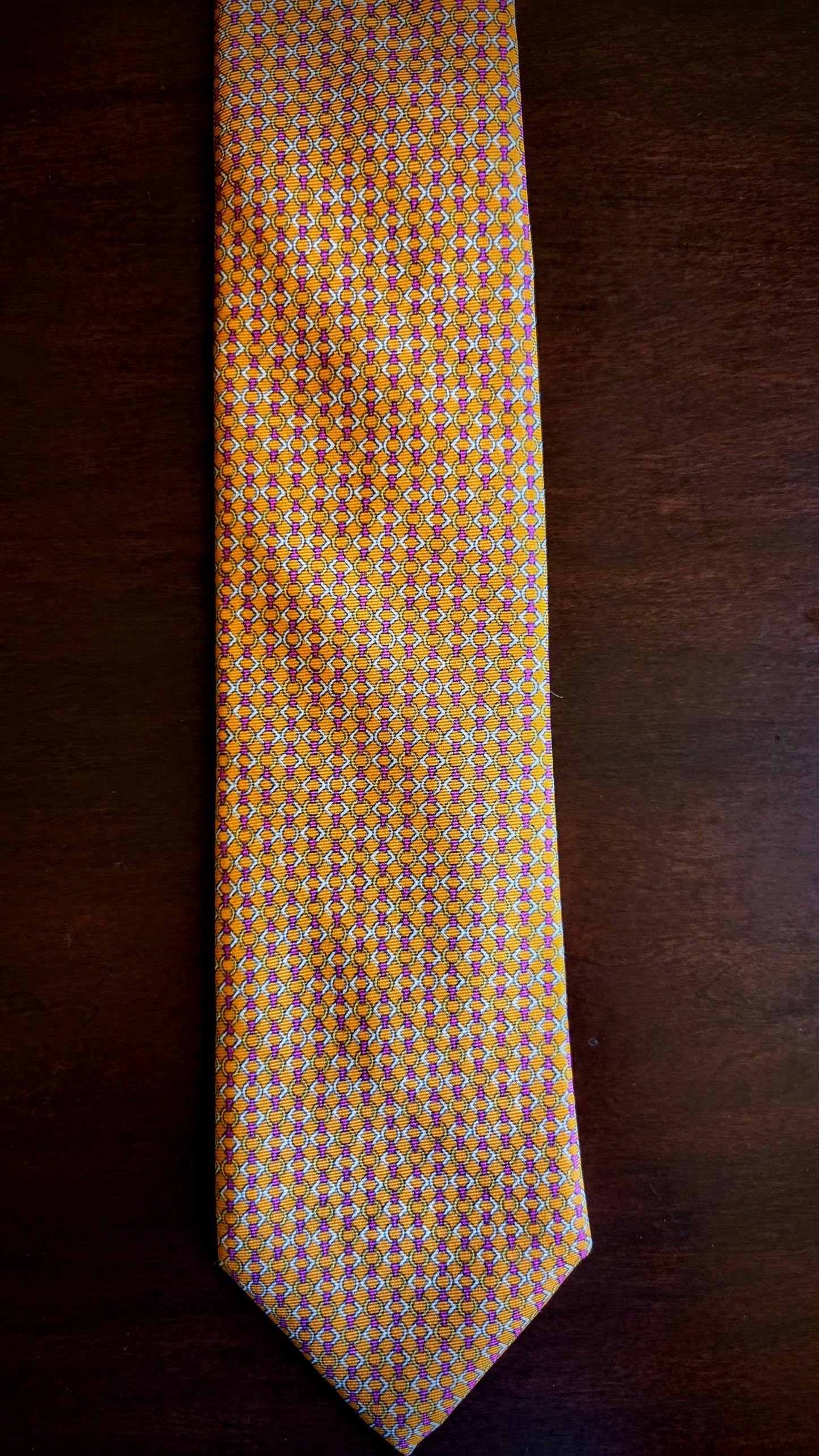 A yellow and orange patterned necktie with small geometric details, laid flat on a dark brown wooden surface.