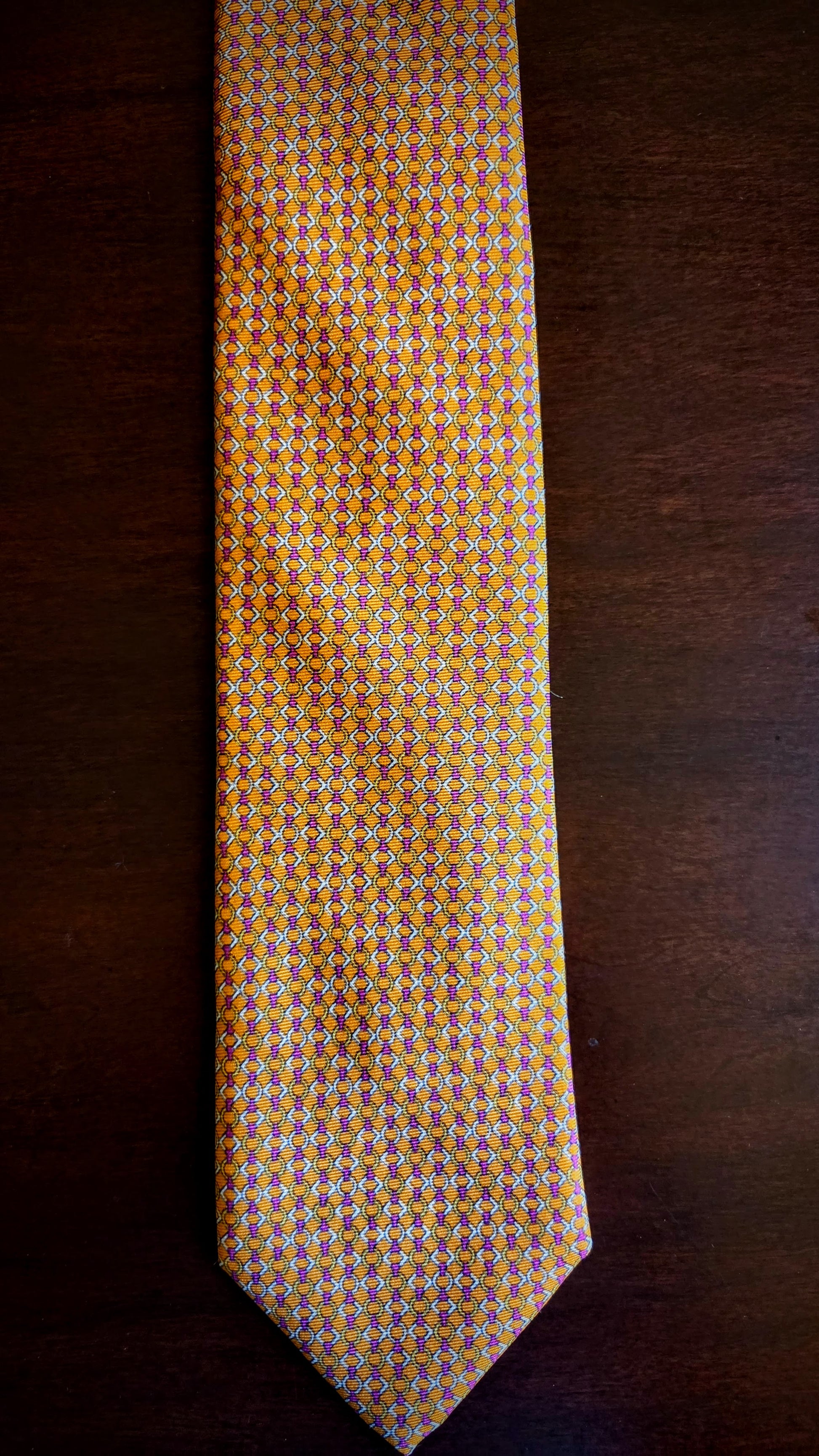 A yellow and orange patterned necktie with small geometric details, laid flat on a dark brown wooden surface.