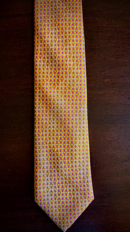 A yellow and orange patterned necktie with small geometric details, laid flat on a dark brown wooden surface.