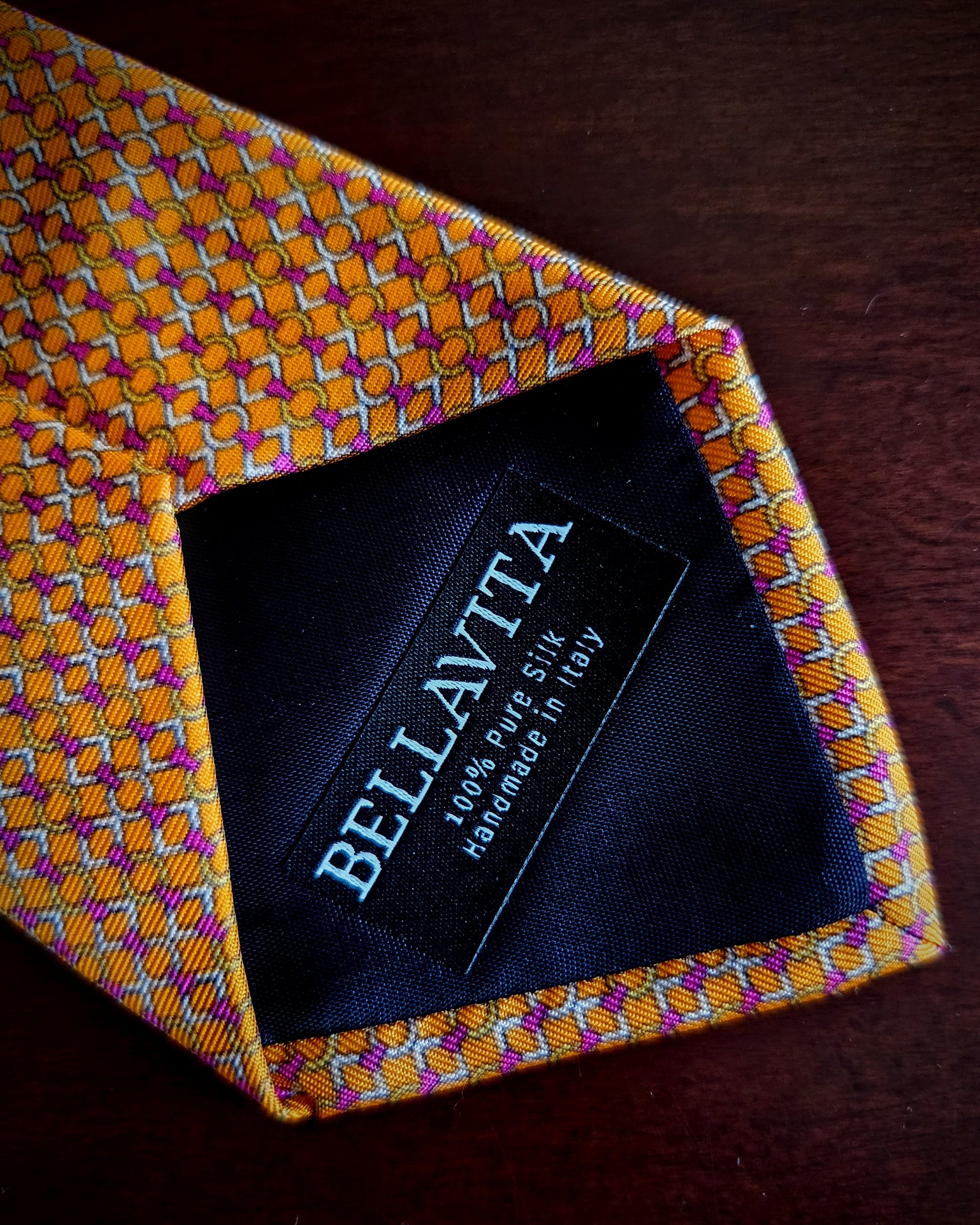 A close-up of a Bellavita silk tie with a yellow, orange, and purple geometric pattern, laid flat on a dark wooden surface. The inside neckband is black with the brand name 'BELLAVITA' printed in white.