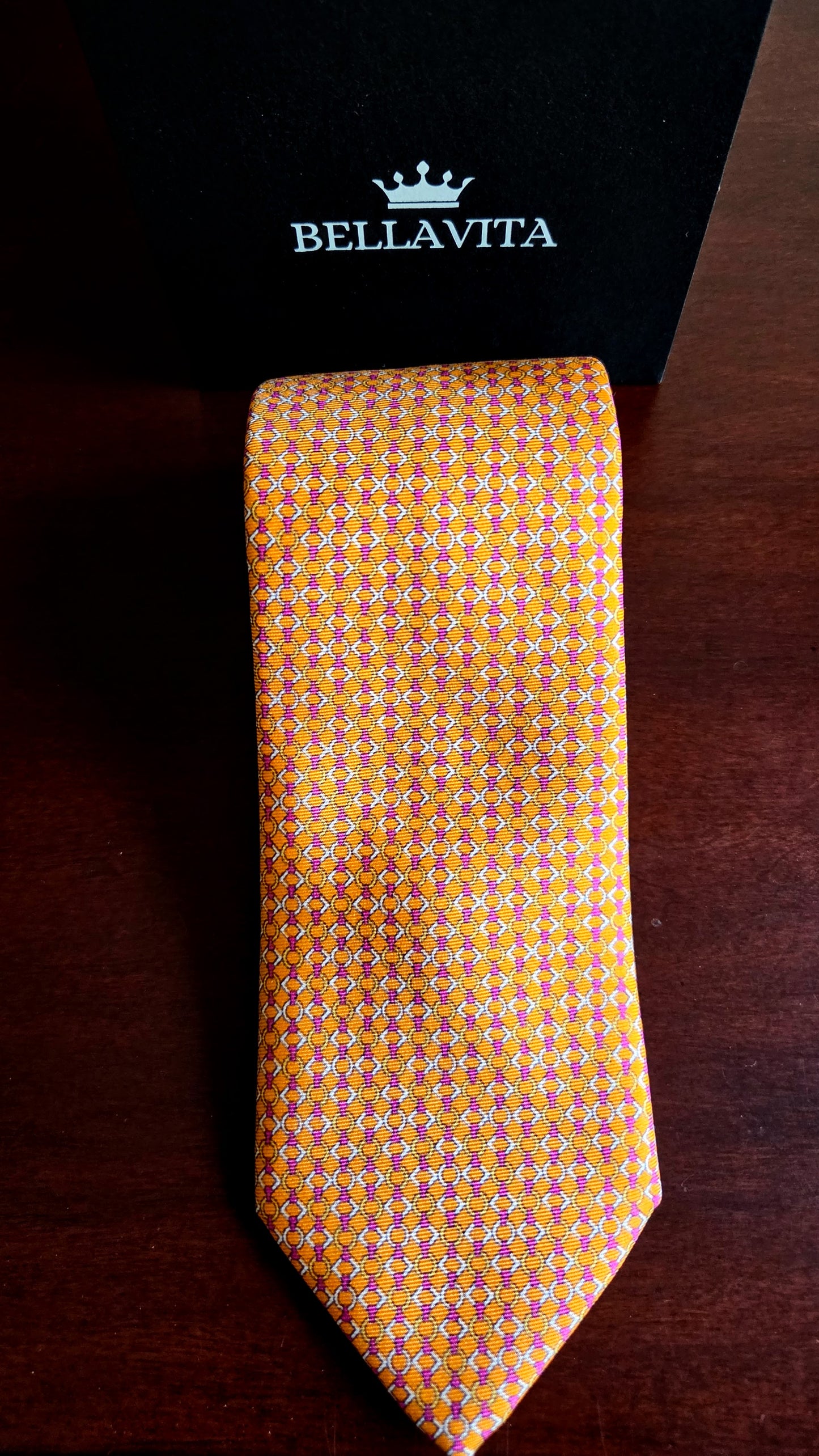 A Bellavita necktie with a yellow and orange geometric pattern, displayed vertically next to its black branded box on a dark wooden surface.