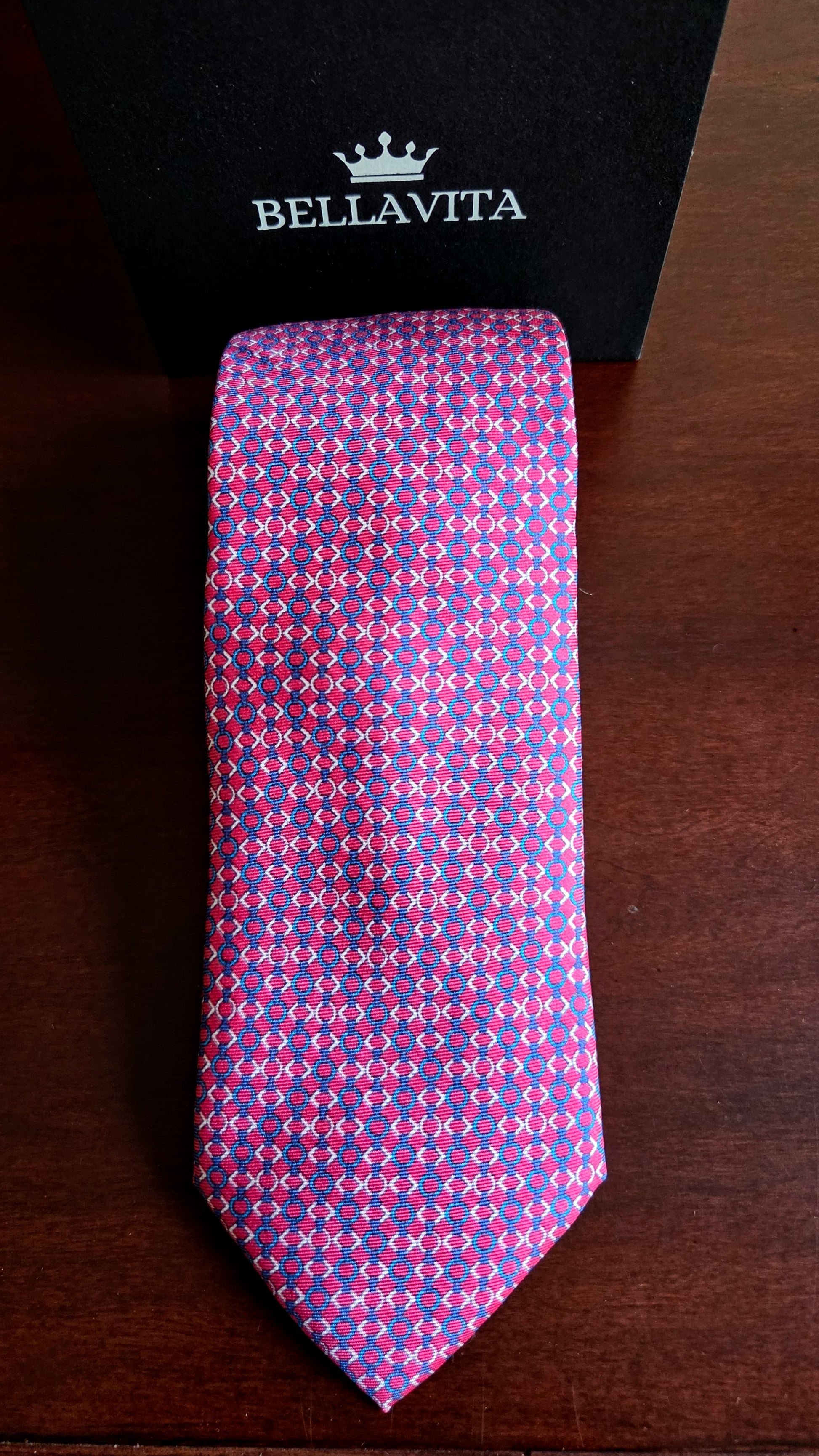 A patterned silk necktie with a purple and pink geometric design, displayed vertically next to its black Bellavita brand box on a dark wooden surface.