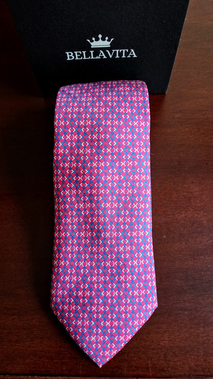 A patterned silk necktie with a purple and pink geometric design, displayed vertically next to its black Bellavita brand box on a dark wooden surface.