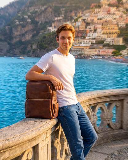 A man in a white t-shirt and blue jeans leans against a stone railing overlooking a scenic coastal town with turquoise water, holding a brown leather backpack.