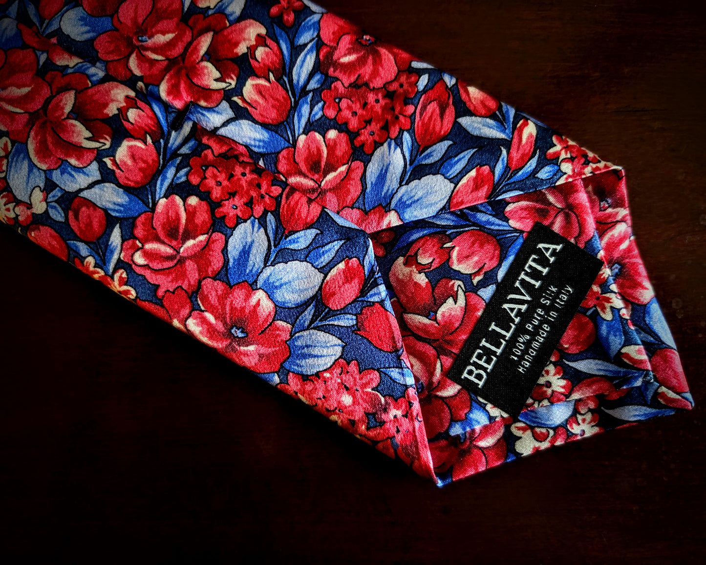 A folded necktie by Bellavita featuring a vibrant floral pattern with red and pink flowers, blue leaves, and white highlights on a dark navy background, displayed on a dark wooden surface.