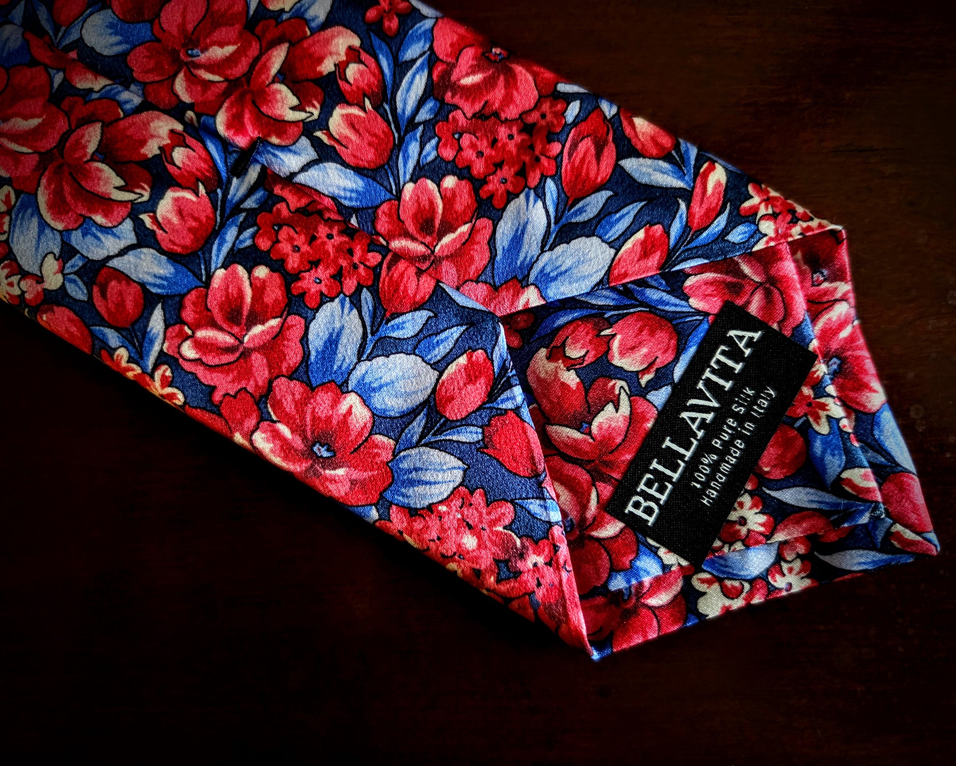 A folded necktie by Bellavita featuring a vibrant floral pattern with red and pink flowers, blue leaves, and white highlights on a dark navy background, displayed on a dark wooden surface.
