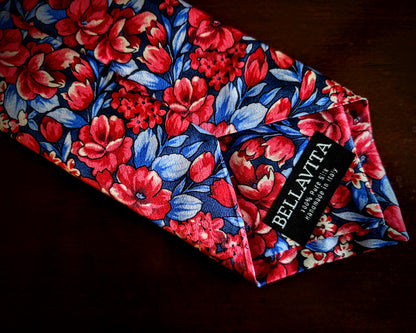 A folded necktie by Bellavita featuring a vibrant floral pattern with red and pink flowers, blue leaves, and white highlights on a dark navy background, displayed on a dark wooden surface.