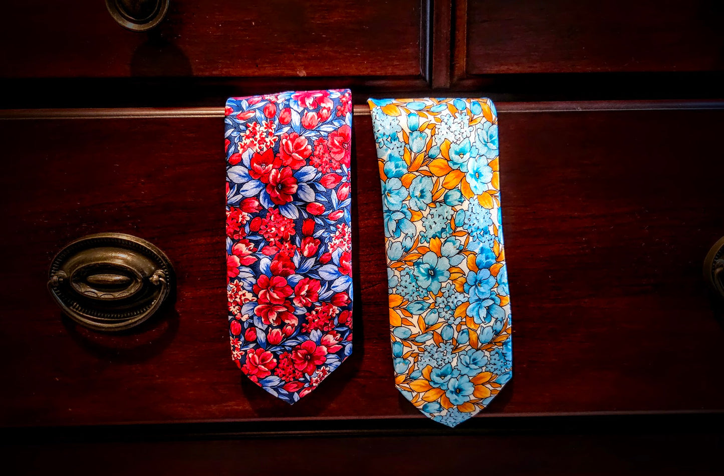 Two patterned neckties displayed side by side on a dark wooden drawer front. The tie on the left has a red and blue floral print, and the tie on the right has a blue and orange floral print.