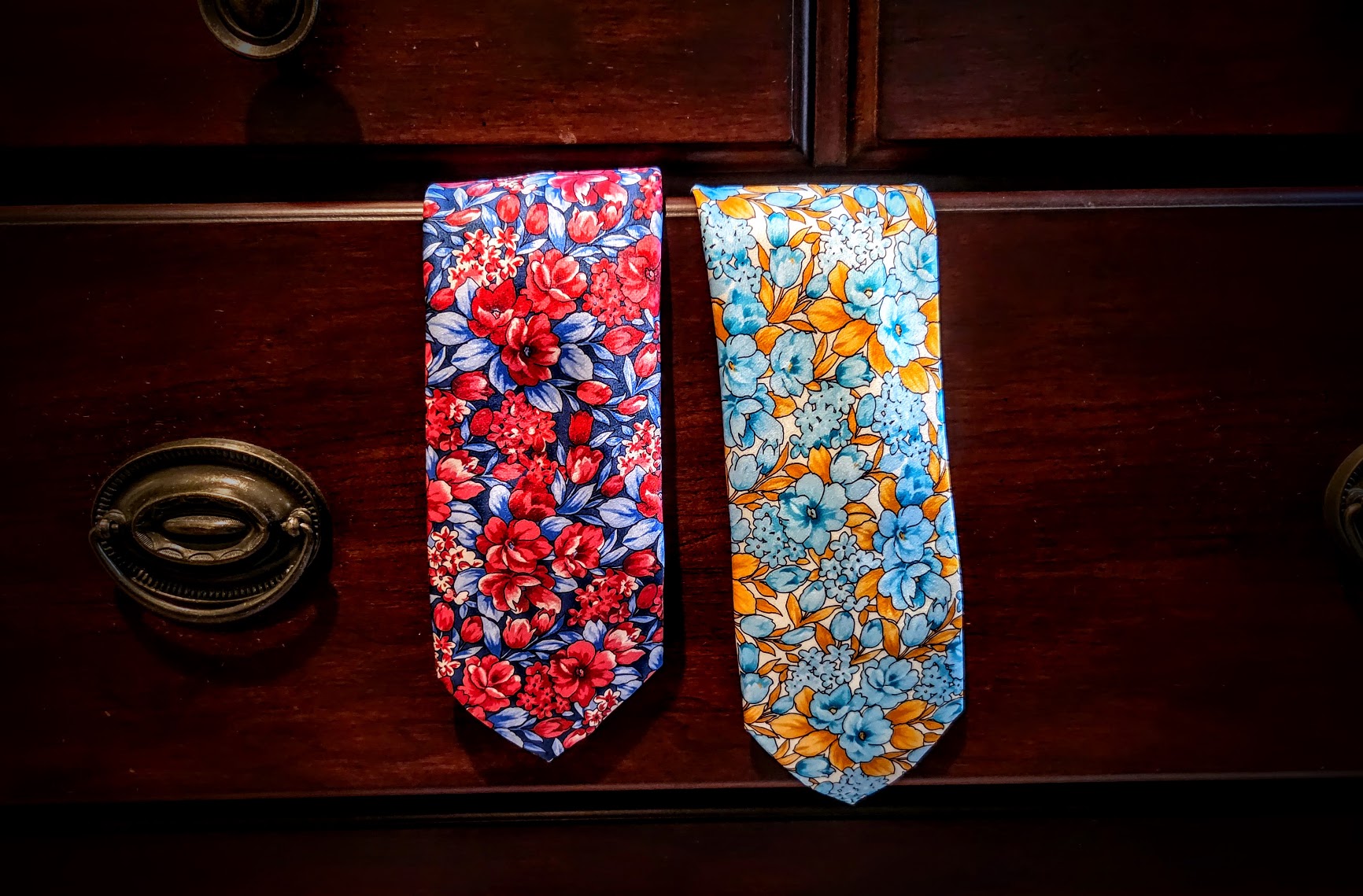 Two patterned neckties displayed side by side on a dark wooden drawer front. The tie on the left has a red and blue floral print, and the tie on the right has a blue and orange floral print.