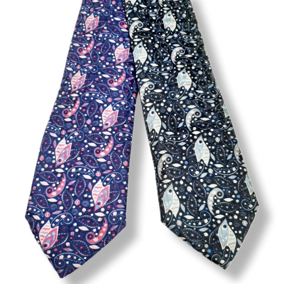 Two patterned neckties displayed side by side on a white background. The left tie is navy blue with pink and white paisley motifs, and the right tie is black with white and light blue paisley motifs.