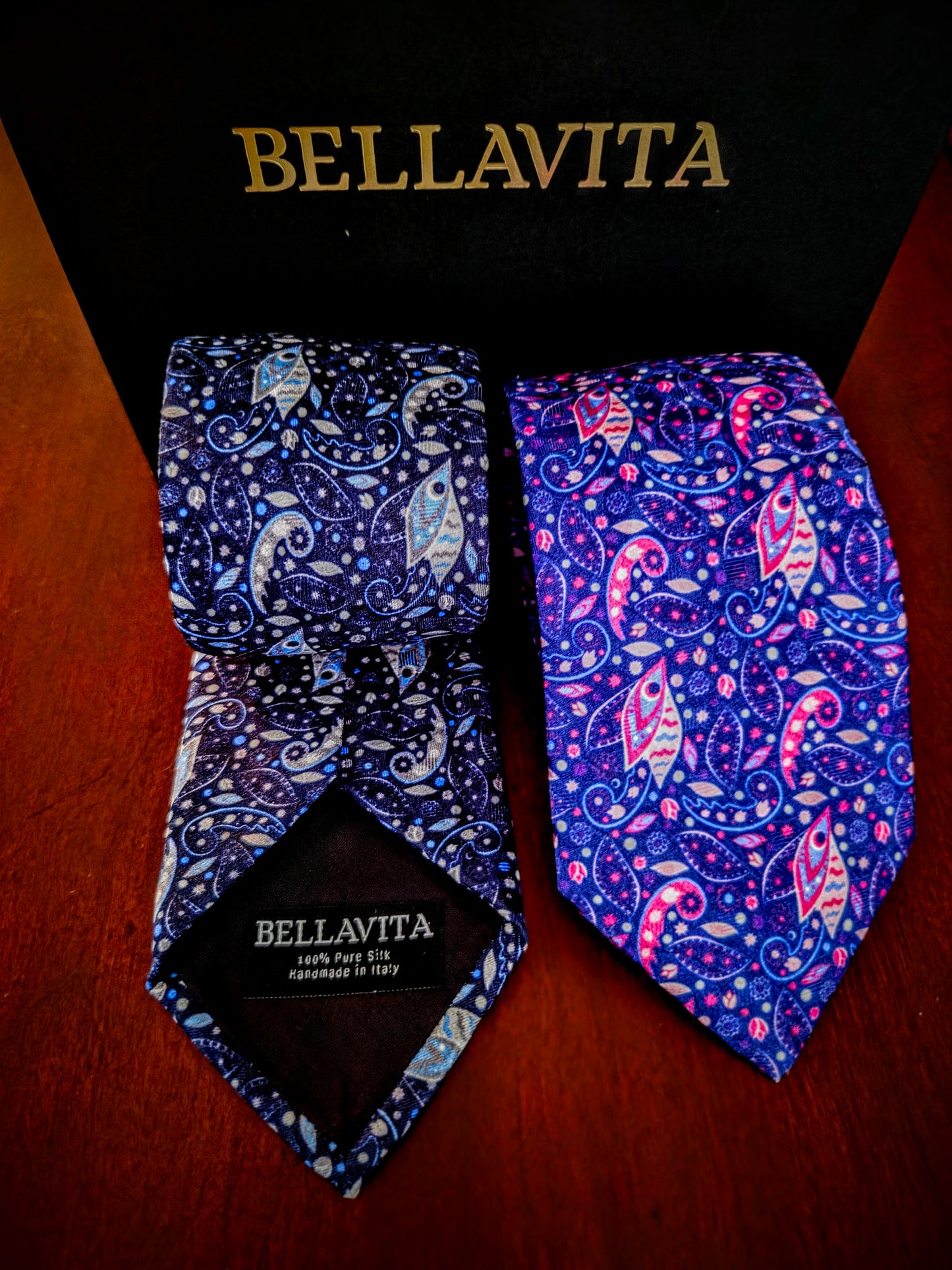 Two Bellavita neckties displayed in front of a black branded box on a wooden surface. One tie is dark blue with white paisley and small pink accents, and the other is a brighter blue-purple with pink, white, and light blue paisley motifs.