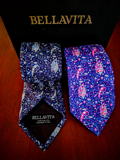 Two Bellavita neckties displayed in front of a black branded box on a wooden surface. One tie is dark blue with white paisley and small pink accents, and the other is a brighter blue-purple with pink, white, and light blue paisley motifs.