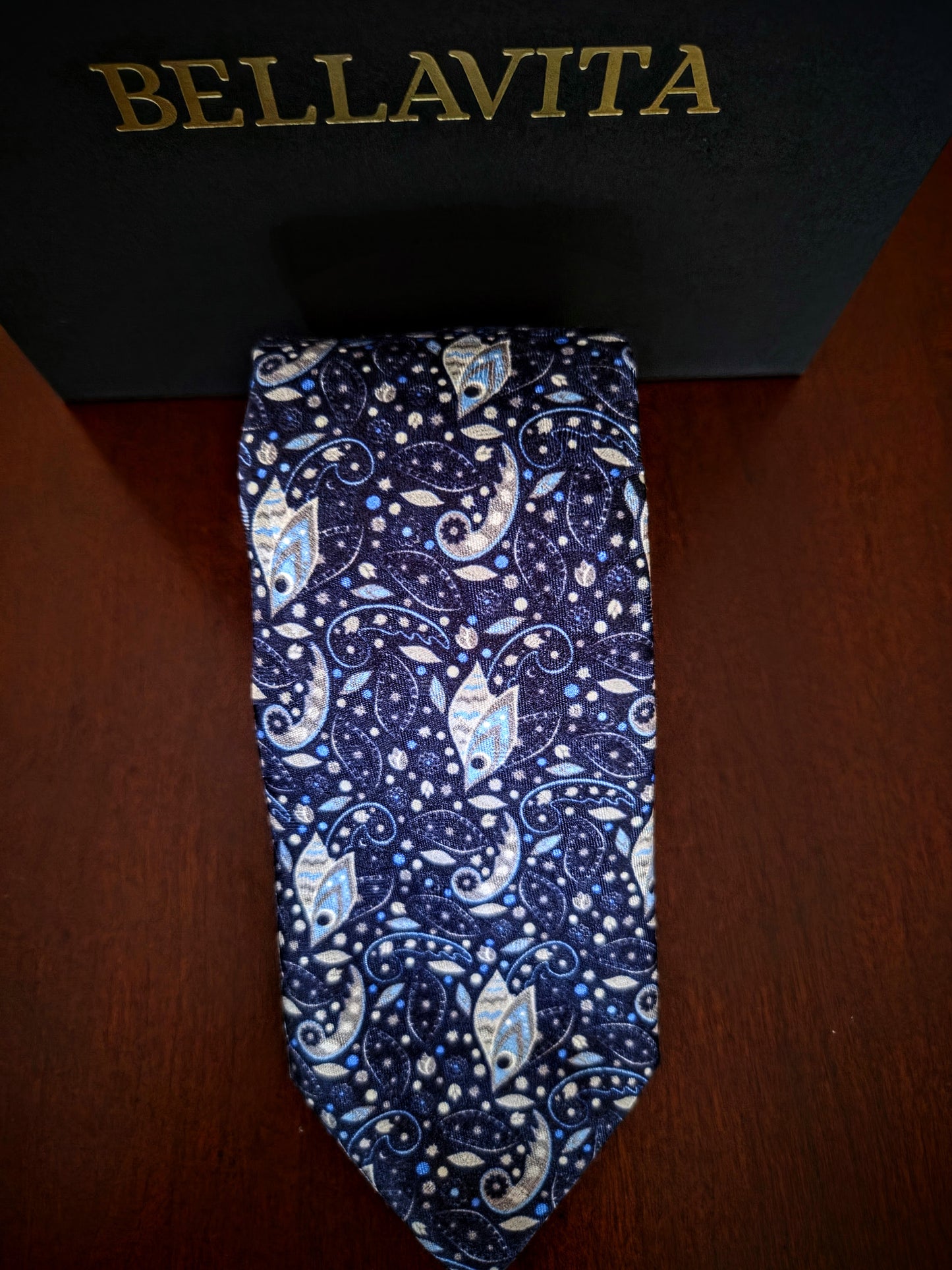 A Bellavita necktie with a blue, white, and light blue paisley pattern, displayed vertically in front of its black branded box on a dark wooden surface.