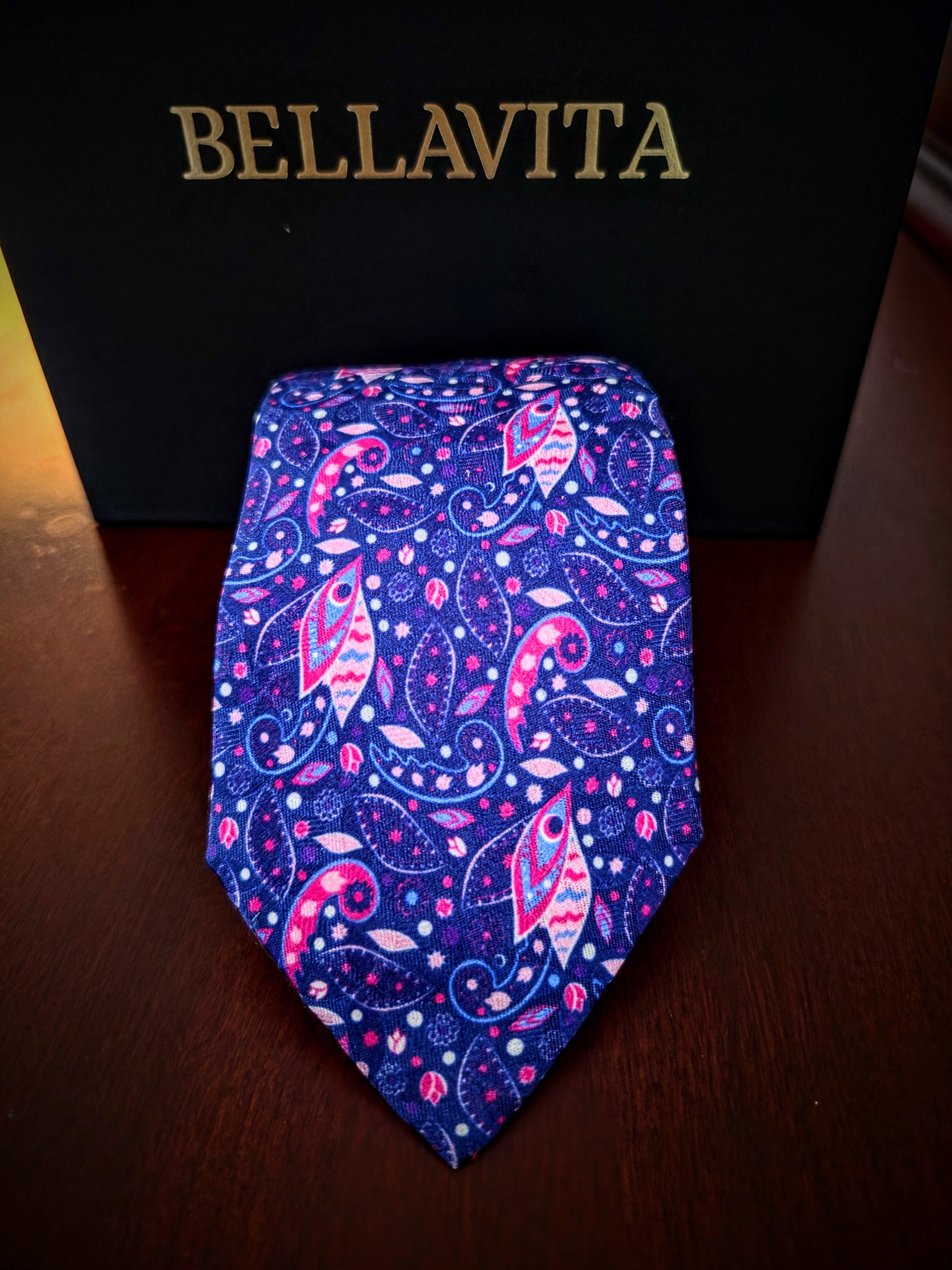 A paisley necktie with a vibrant blue, pink, and purple pattern, displayed in front of a black Bellavita box on a dark wooden surface.