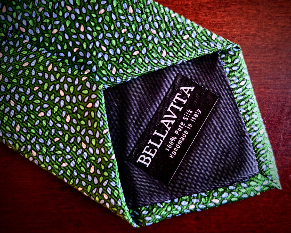 A close-up of a green patterned necktie with a black fabric label reading BELLAVITA, placed on a dark reddish-brown wooden surface.