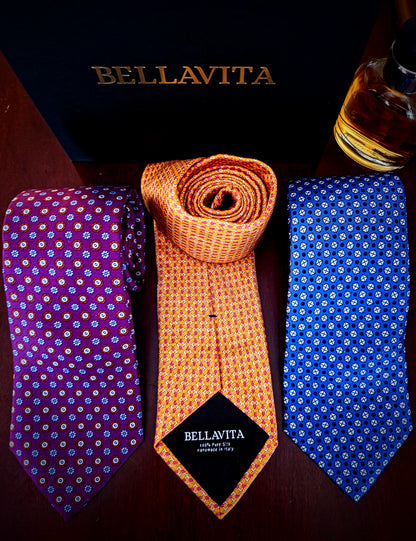 Three Bellavita silk neckties displayed on a dark wooden surface with the brand's black box in the background. The left tie is purple with a white and orange floral pattern, the center tie is orange with a yellow geometric pattern and a black brand label, and the right tie is blue with a white floral pattern.