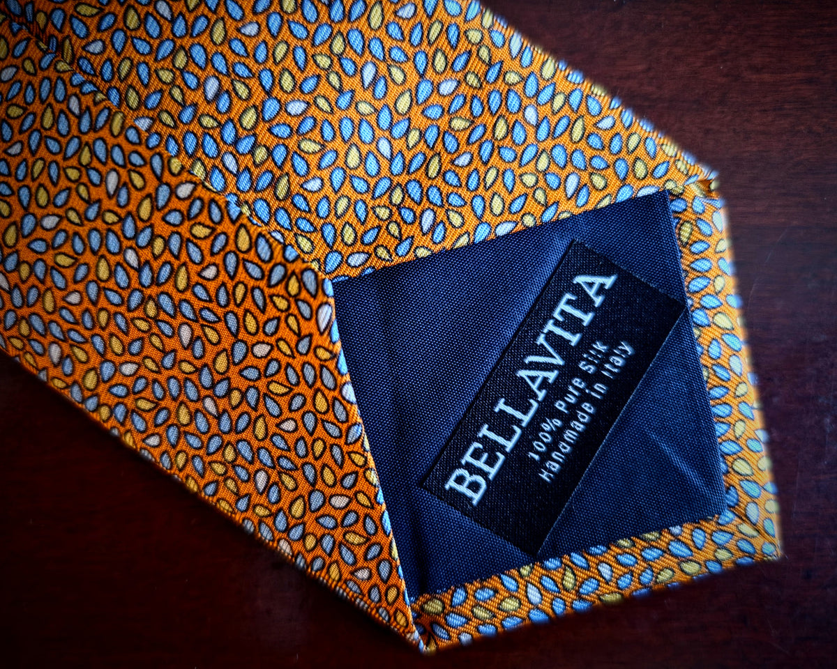 A close-up of a Bellavita silk necktie with a vibrant orange and blue geometric pattern, laid flat on a dark wooden surface. The inside collar features a dark navy blue fabric lining with a black brand label.