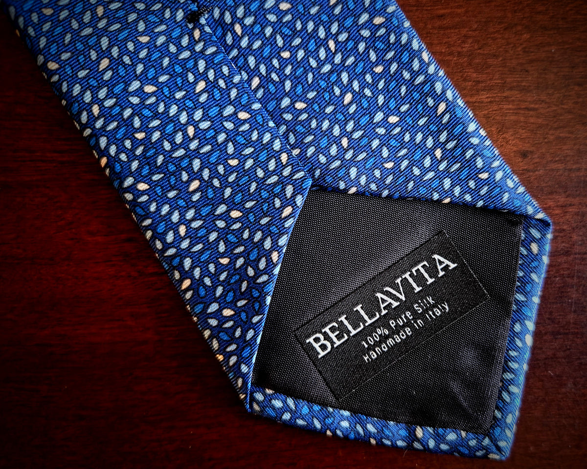 A close-up of a blue patterned necktie with a black Bellavita brand label, laid on a dark wooden surface.