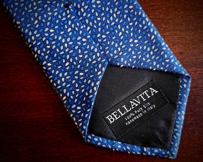A close-up of a blue patterned necktie with a black Bellavita brand label, laid on a dark wooden surface.