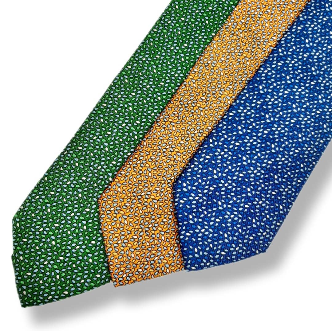 Three patterned neckties arranged diagonally on a white background, featuring green, mustard yellow, and blue micro-dot prints.