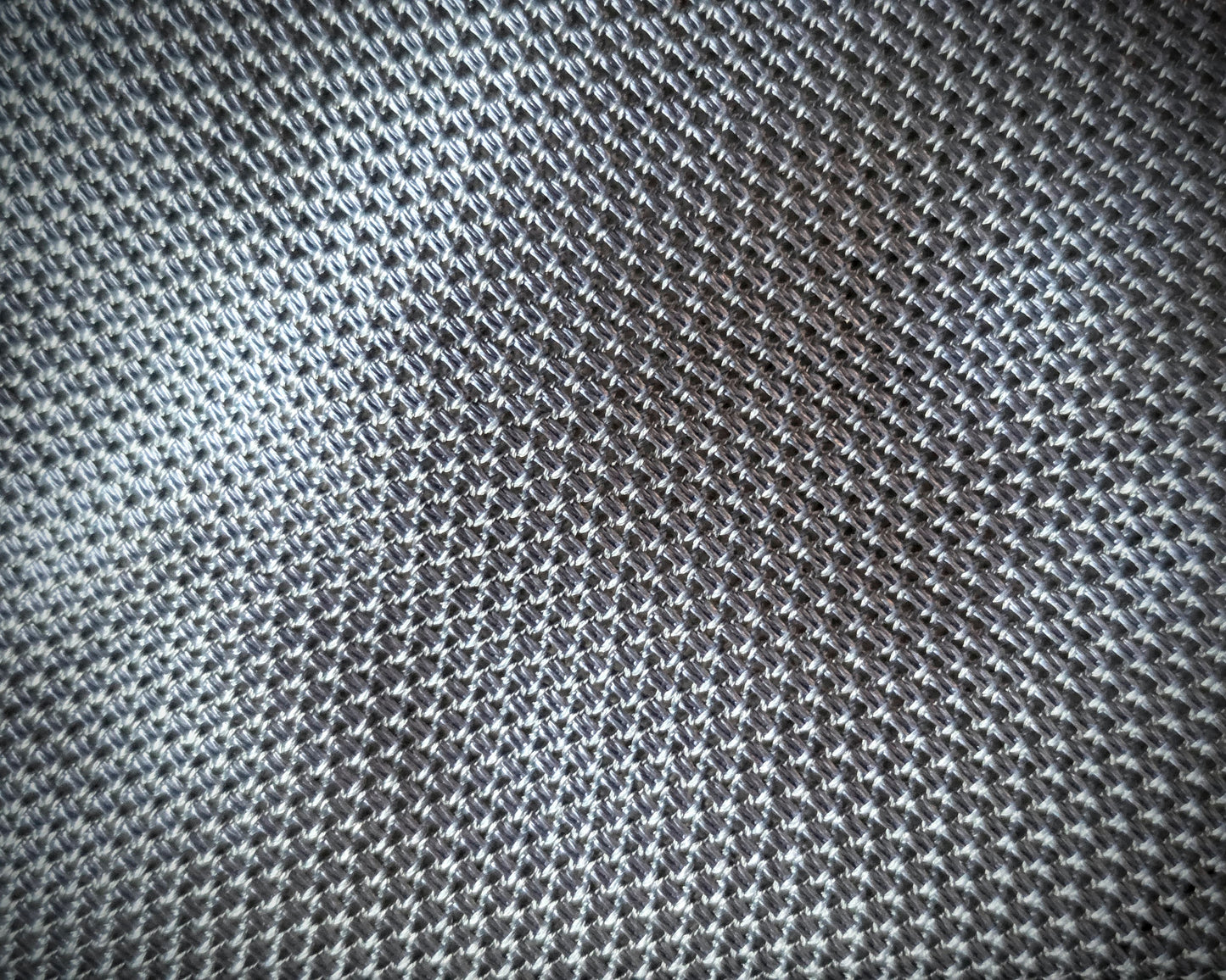 A close-up of a textured fabric with a fine woven pattern in gray and white, showing the material's details and subtle sheen.