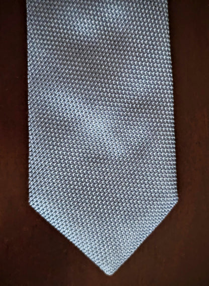 A textured silver necktie with a fine woven pattern, laid flat on a dark brown wooden surface.