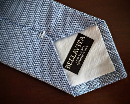 A close-up of a light blue and white textured silk tie with a black and white Bellavita brand label sewn inside the tip, resting on a dark brown wooden surface.