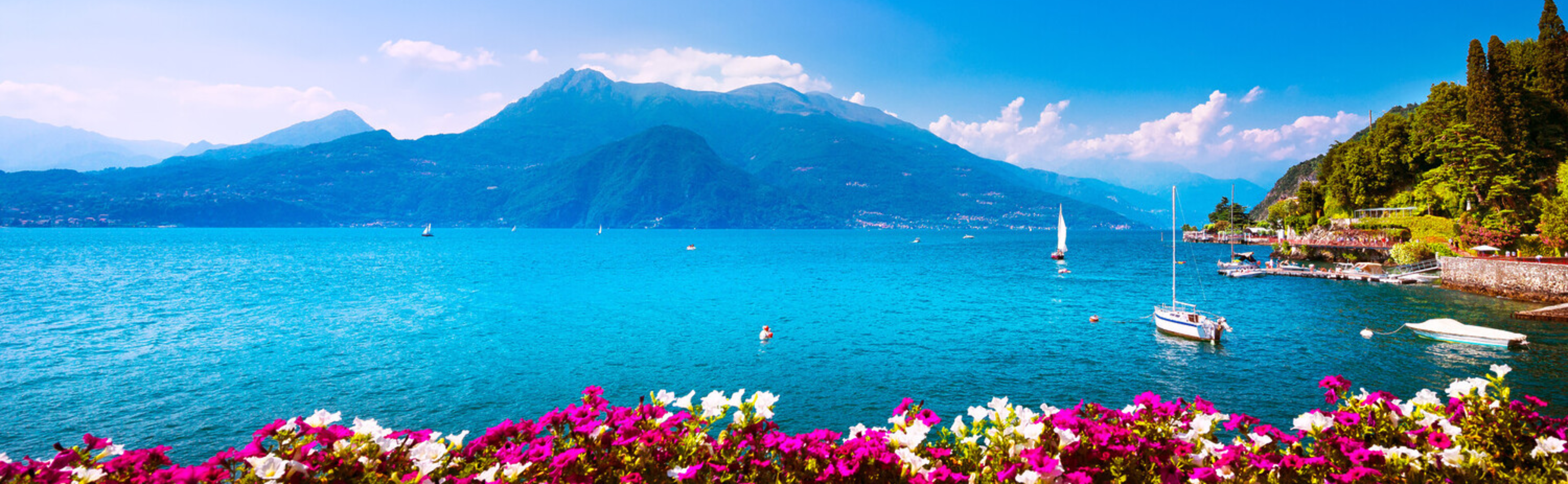 A vibrant scenic landscape print of a mountain lake, featuring bright turquoise water, sailboats, a snow-capped mountain in the background, and a foreground of colorful pink and white flowers.