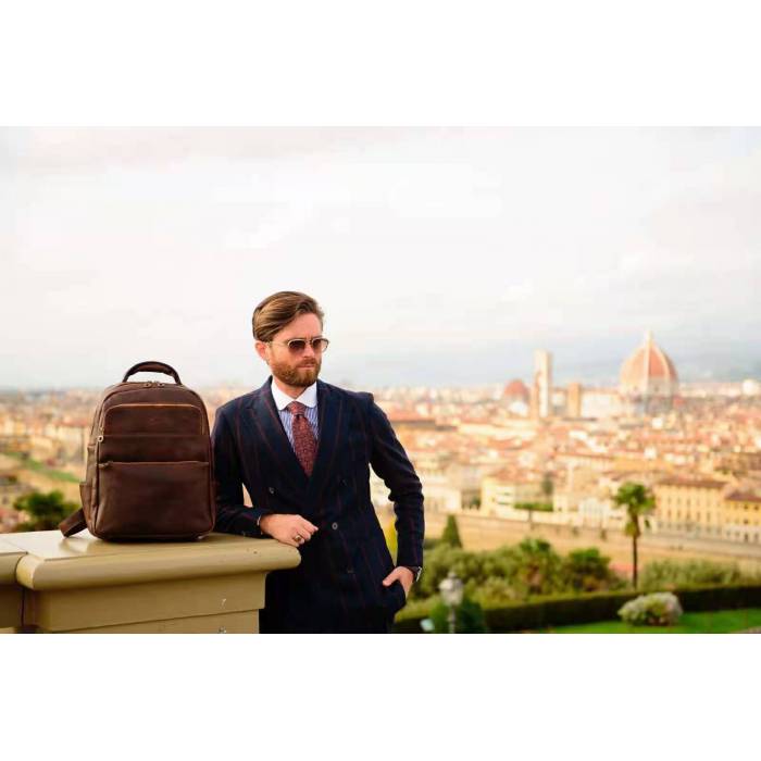 A man in a dark navy suit stands outdoors next to a brown leather backpack on a stone ledge, with a panoramic cityscape in the background.