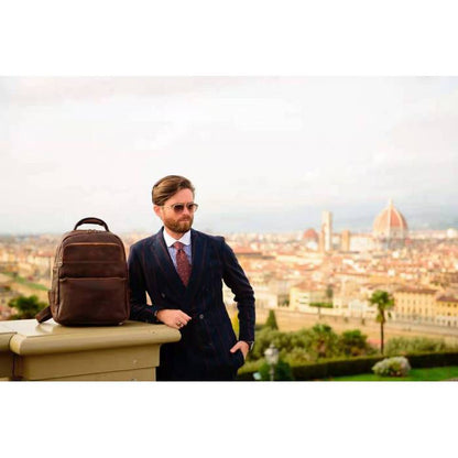 A man in a dark navy suit stands outdoors next to a brown leather backpack on a stone ledge, with a panoramic cityscape in the background.