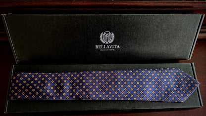 A blue patterned necktie with small gold and white motifs, presented inside an open black velvet Bellavita gift box on a dark wooden surface.
