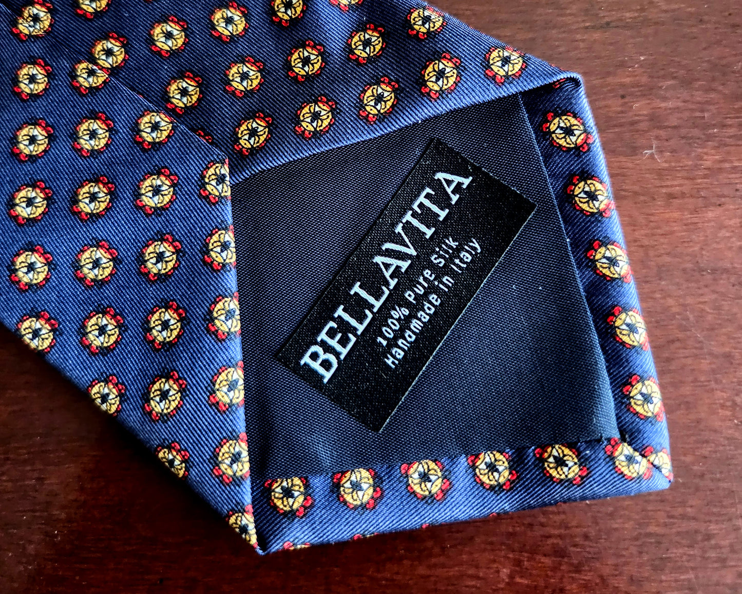 A close-up of a Bellavita necktie on a dark wooden surface, showing a blue base with a repeating yellow, red, and white floral pattern and a black brand label.