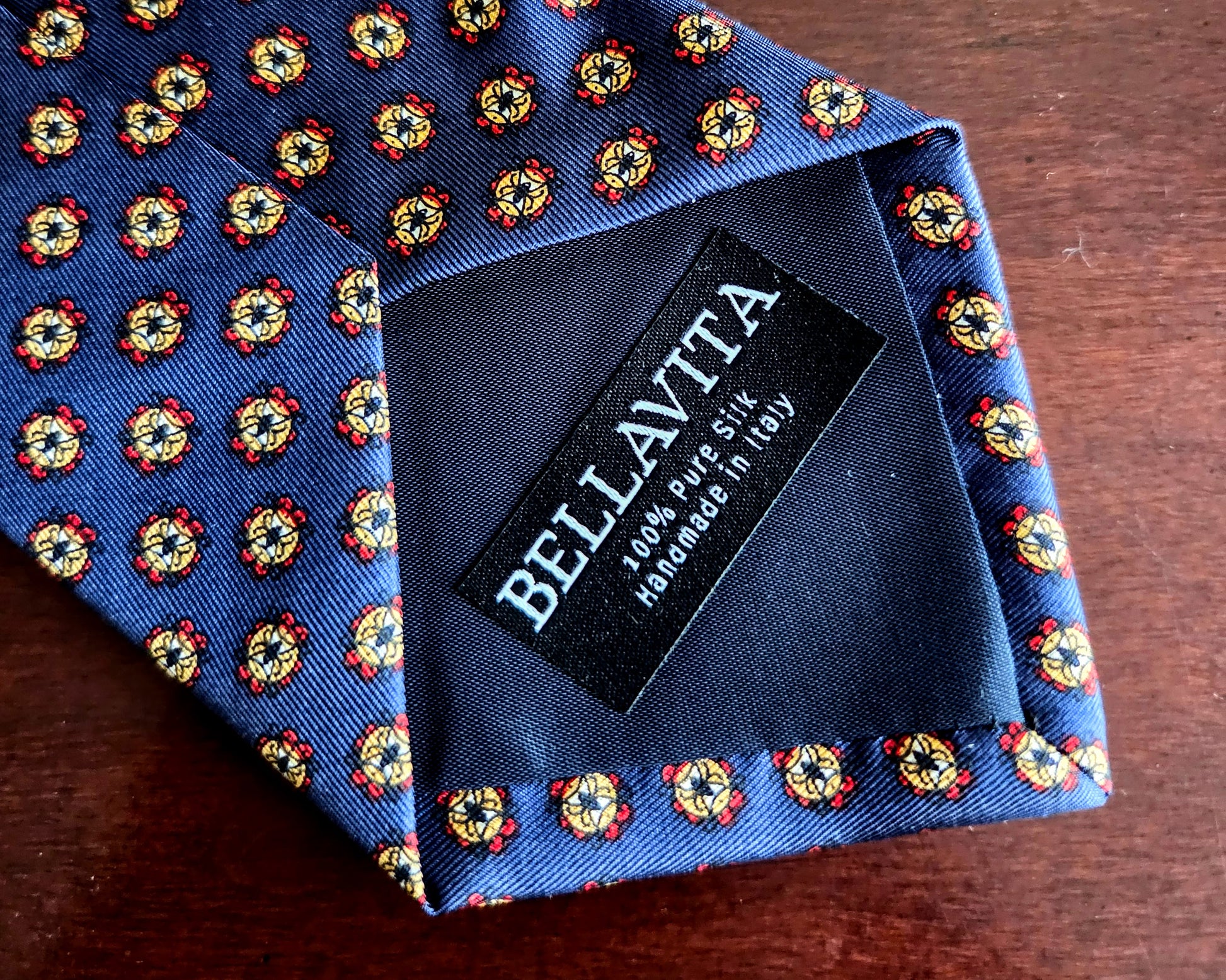 A close-up of a Bellavita necktie on a dark wooden surface, showing a blue base with a repeating yellow, red, and white floral pattern and a black brand label.