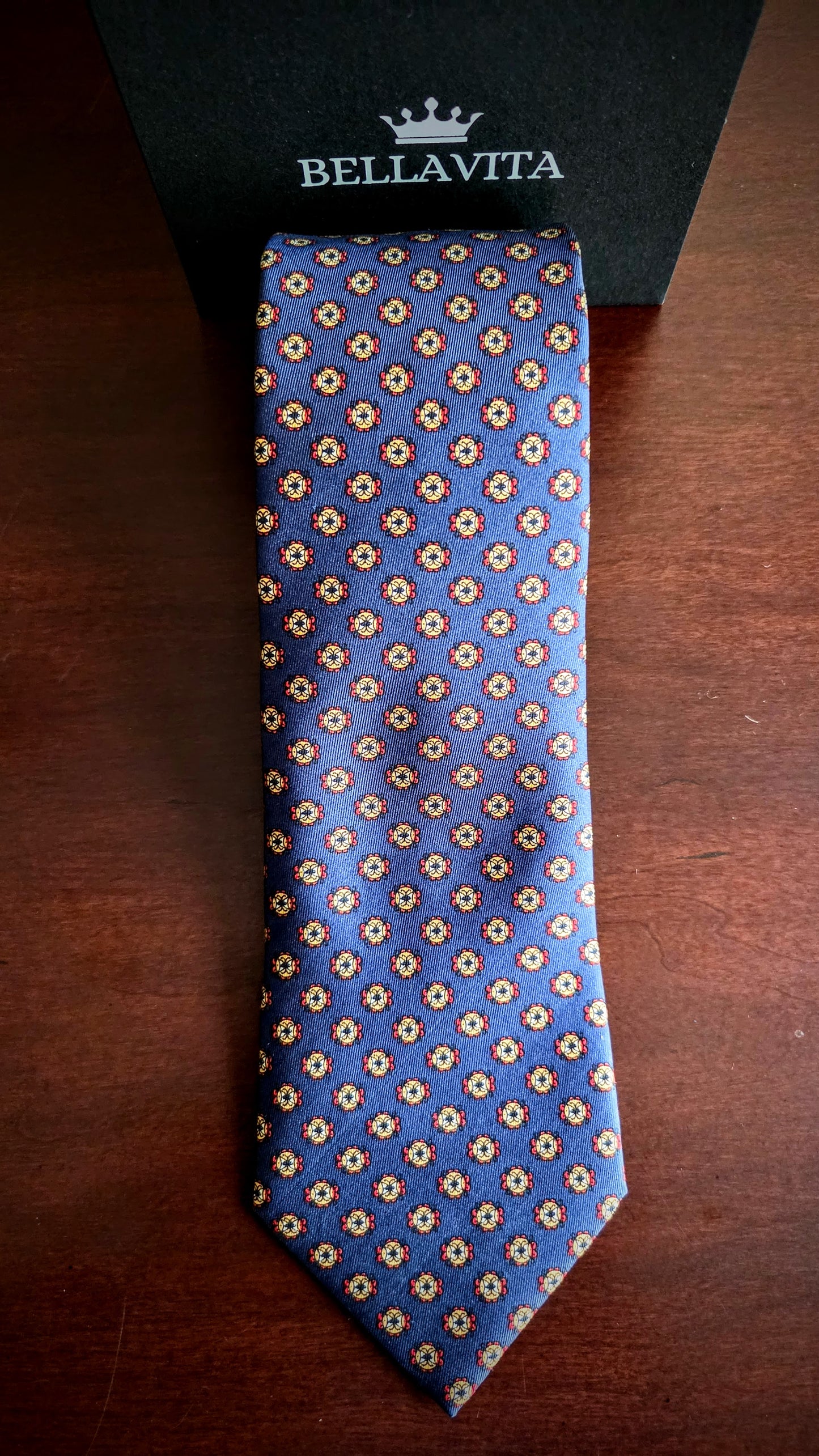 A blue silk necktie with a repeating beige and gold floral pattern, laid flat on a dark wooden surface next to its black Bellavita brand box.