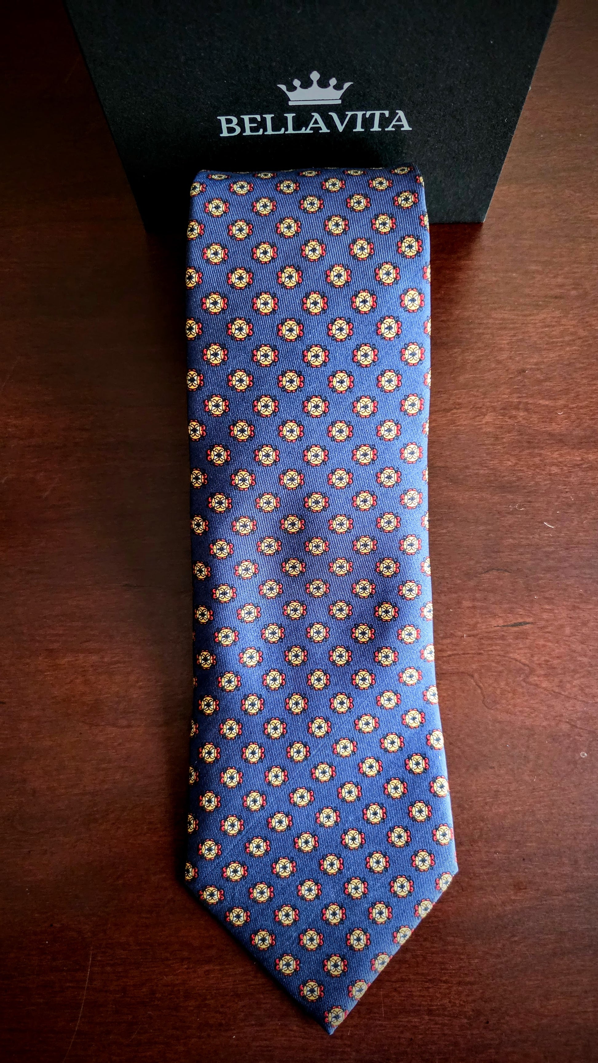 A blue silk necktie with a repeating beige and gold floral pattern, laid flat on a dark wooden surface next to its black Bellavita brand box.
