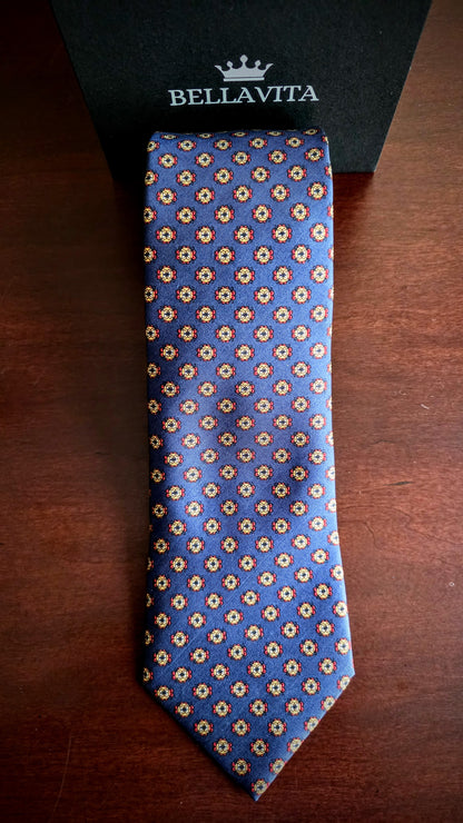 A blue silk necktie with a repeating beige and gold floral pattern, laid flat on a dark wooden surface next to its black Bellavita brand box.