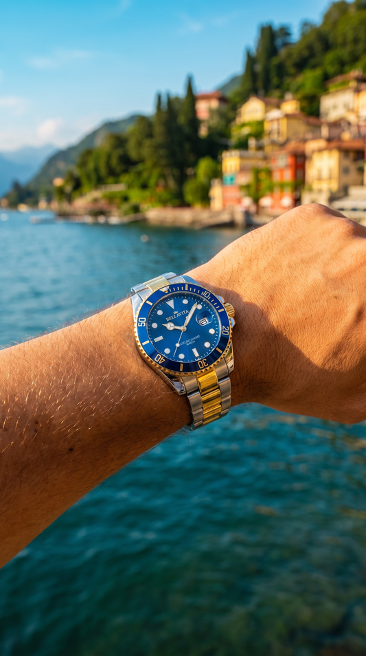 A close-up of a wrist wearing a blue and gold-tone stainless steel watch, with a scenic background of a lake, mountains, and a coastal town.