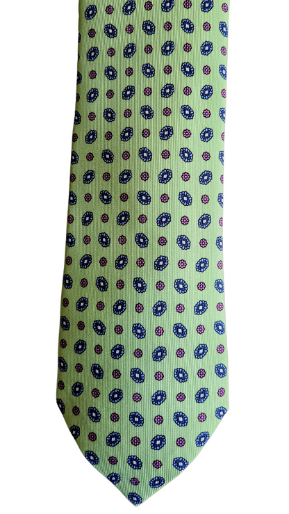 A slim necktie with a light sage green base, featuring a repeating pattern of small blue and purple circular medallions, shown against a plain white background.