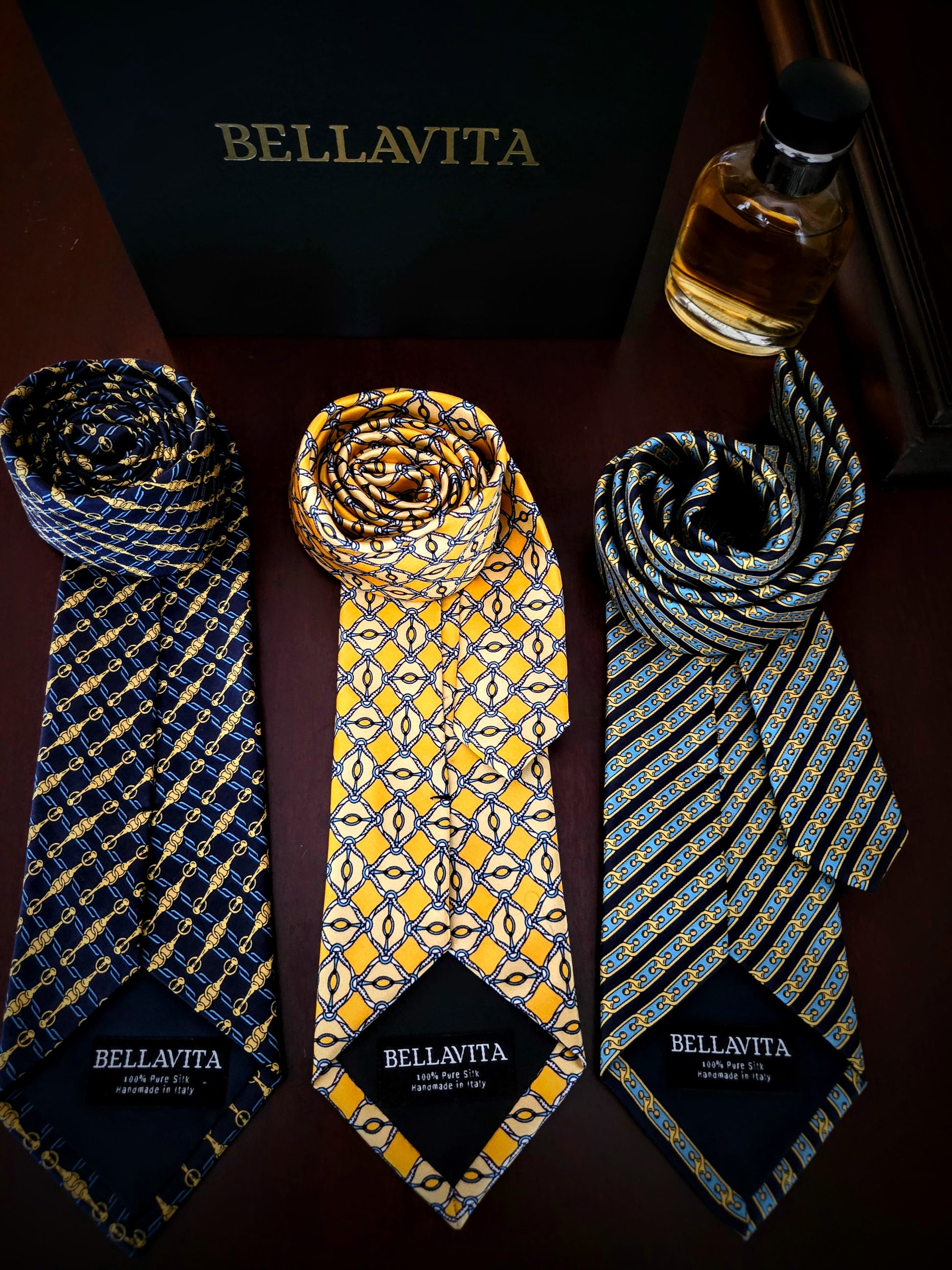 Three Bellavita patterned neckties displayed on a dark surface with a black Bellavita box behind them and a small bottle at the top right. The ties have blue, yellow, and black geometric patterns, each with a black Bellavita label at the bottom.