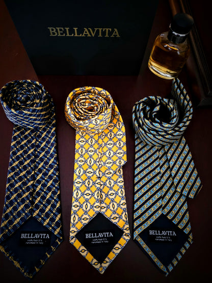Three Bellavita patterned neckties displayed on a dark surface with a black Bellavita box behind them and a small bottle at the top right. The ties have blue, yellow, and black geometric patterns, each with a black Bellavita label at the bottom.